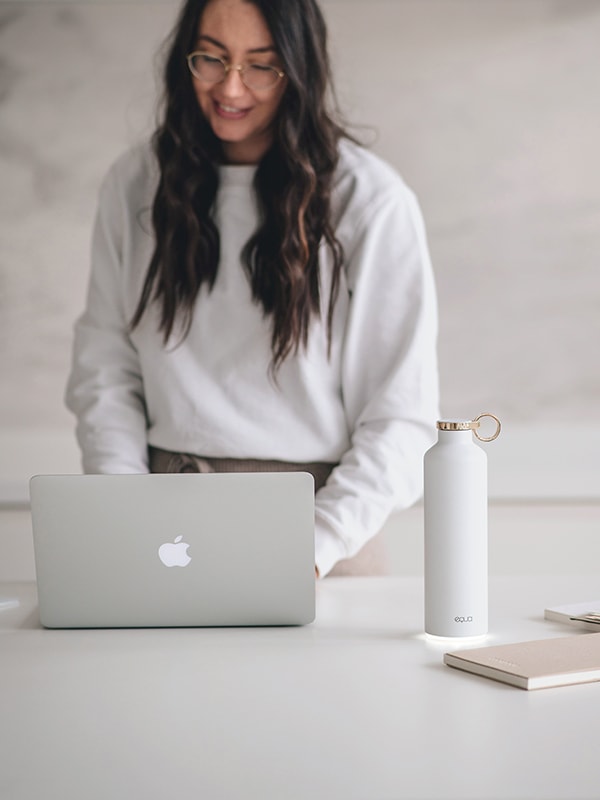 Smart Water Bottle EQUA in White Colour on the right bottom part of the image. With lady in the background working on her lap top. Glow reminder keeping her water intake and hydration always in sync with her needs.